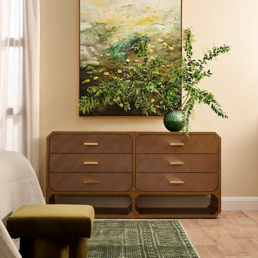 Wooden dresser with gold handles in a room with a painting and plant on the wall.