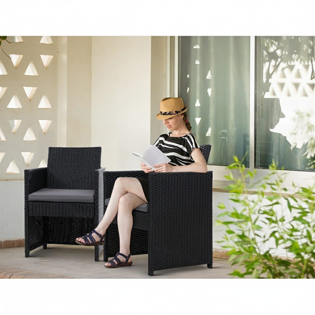 Woman sitting on a black wicker chair reading a book on a patio.